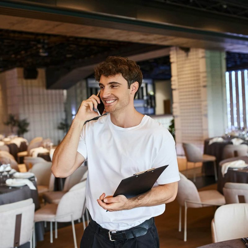 smiling-event-organizer-with-clipboard-talking-on-smartphone-near-festive-tables-in-banquet-hall.jpg